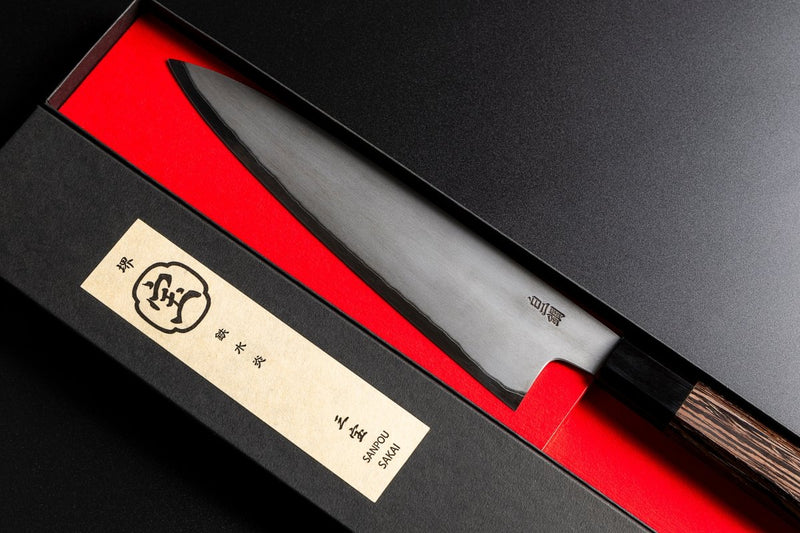 Close-up of the blade of Sakai TAKAYUKI Gyuto 210mm by Aoki Hamono inside its box next to the cover with a Japanese characters label in the cover