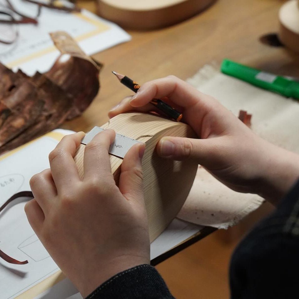 Closeup of hands with a pencil and a small rule measuring up parts for a bento box  with wood scratches in the background inside Hakata Bentwood Tamaki workshop
