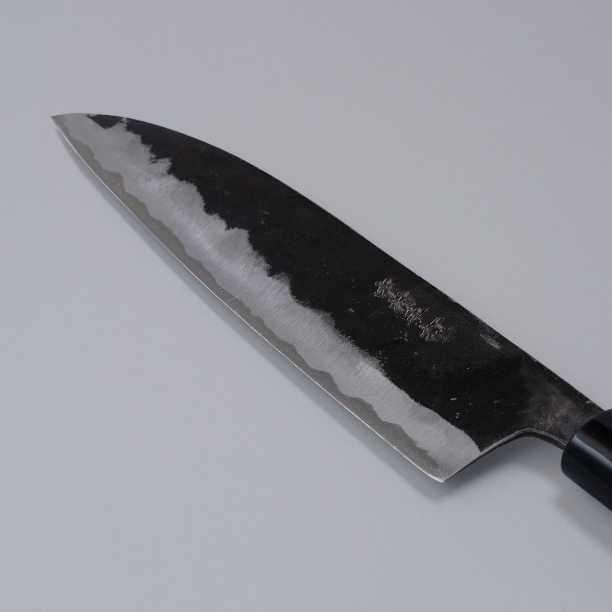 Closeup on engraved blade of Kurouchi Santoku Knife 180mm crafted by Nakamura Blacksmith displayed diagonally on grey background
