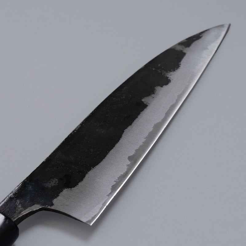 Closeup on blade of Kurouchi Santoku Knife 150mm crafted by Nakamura Blacksmith displayed diagonally on grey background