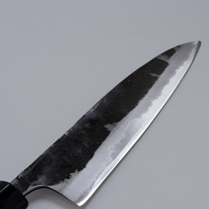 Closeup on blade of Kurouchi Santoku Knife 165mm crafted by Nakamura Blacksmith displayed diagonally on grey background