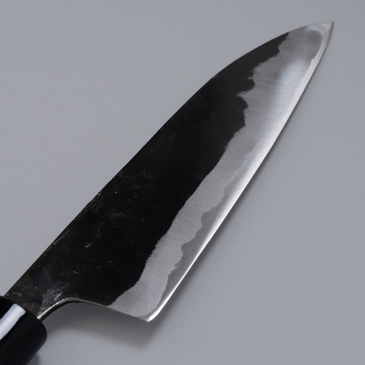 Closeup on reverse side of Kurouchi Santoku Knife 180mm crafted by Nakamura Blacksmith displayed diagonally on grey background