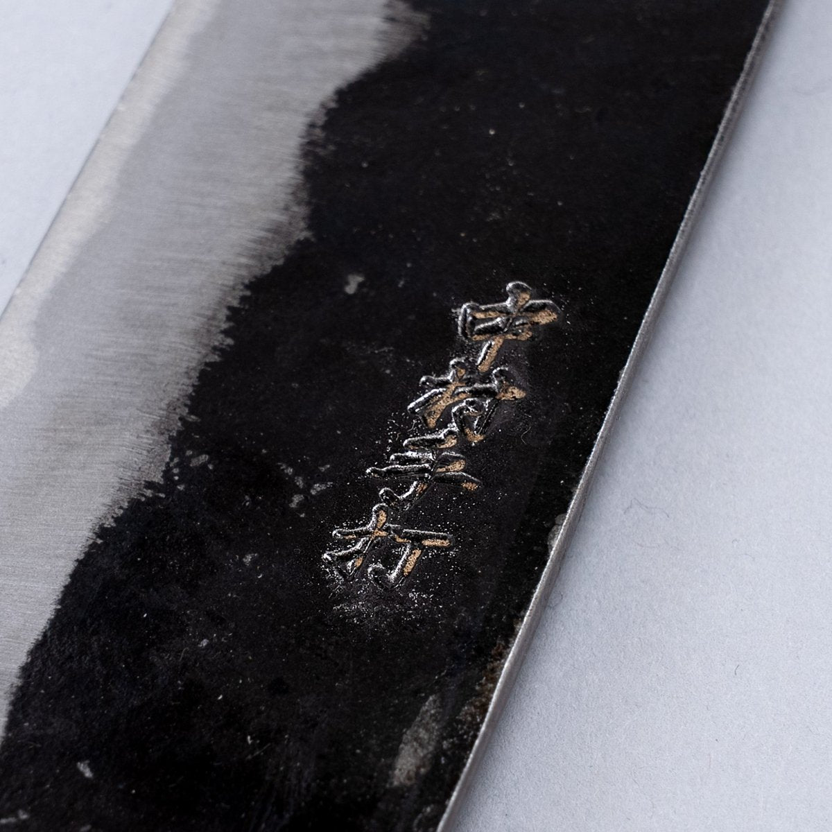Closeup on engraving on Kurouchi Santoku Knife 180mm crafted by Nakamura Blacksmith on grey background