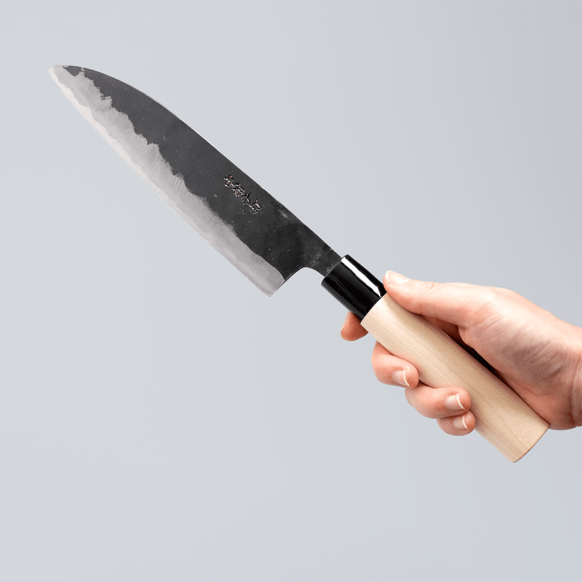 Hand holding Kurouchi Santoku Knife 180mm crafted by Nakamura Blacksmith on grey background