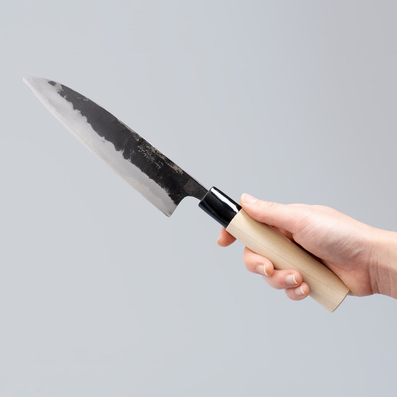 Hand holding Kurouchi Santoku Knife 165mm crafted by Nakamura Blacksmith on grey background