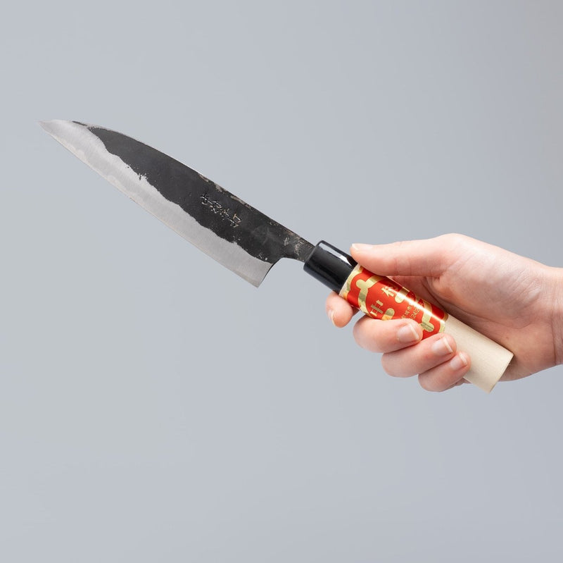 Hand holding Kurouchi Santoku Knife 150mm crafted by Nakamura Blacksmith  on grey background