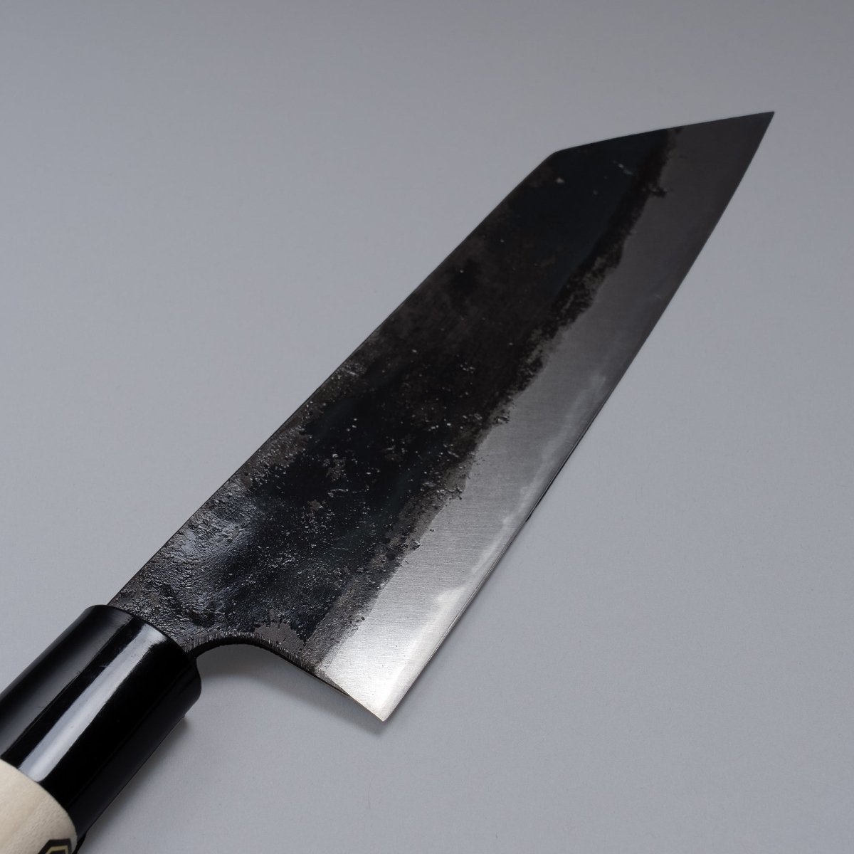 Closeup on blade of Kurouchi Santoku knife 160mm crafted by Shigemitsu Hamono displayed diagonally on grey background 
