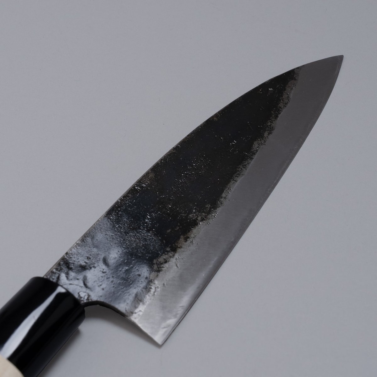 Closeup on blade of Kurouchi Deba knife 155mm crafted by Shigemitsu Hamono displayed diagonally on grey background 