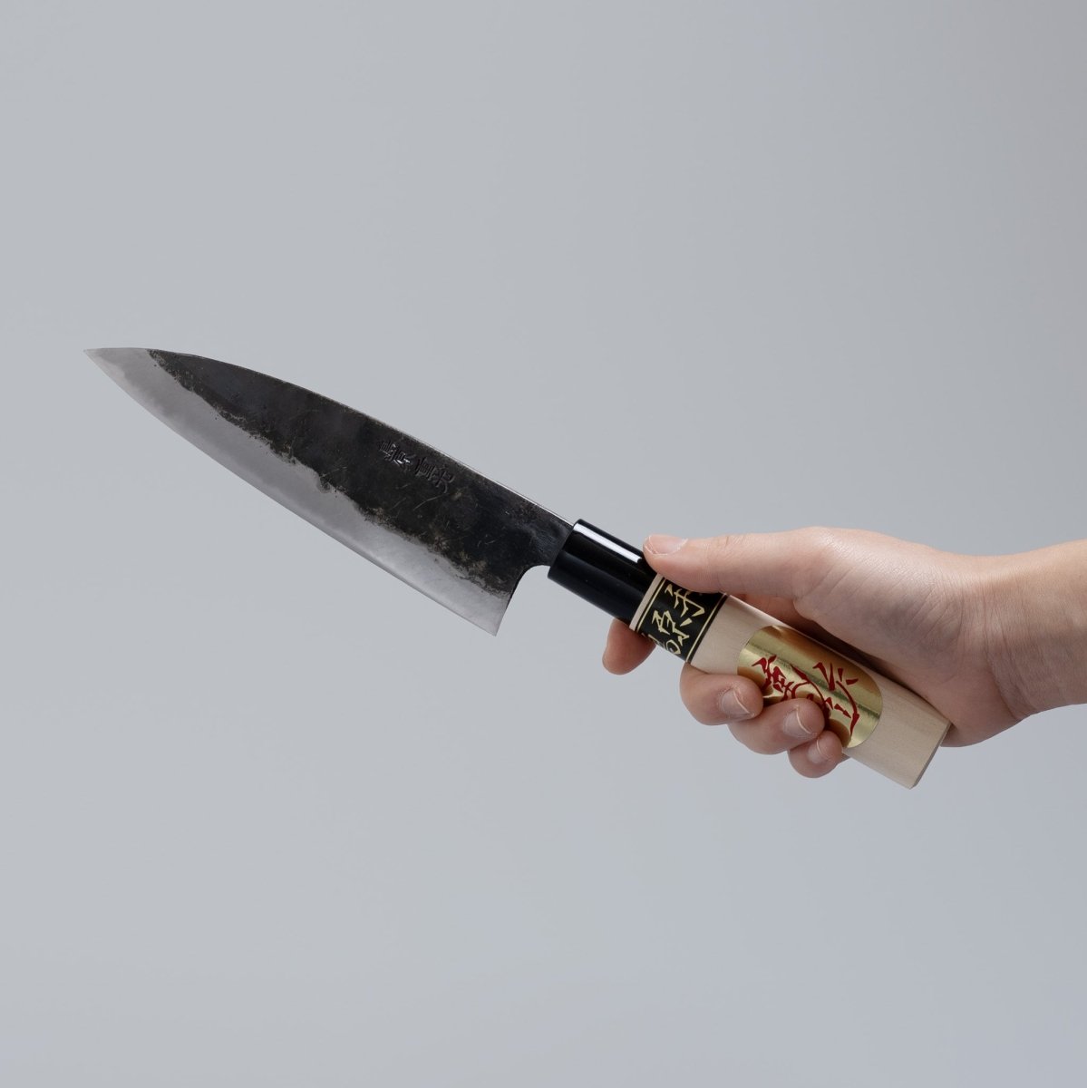 Hand holding Kurouchi Deba knife 155mm crafted by Shigemitsu Hamono on grey background 