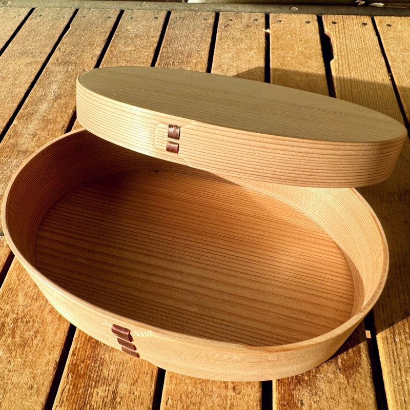 Empty Oval Bento Box crafted by Hakata Bentwood Tamaki open with lid resting atop it on wooden background outdoors