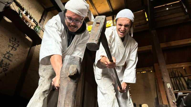Two of the artisan apprentices at Asano Blacksmith in traditional white clothes manually hammering metal
