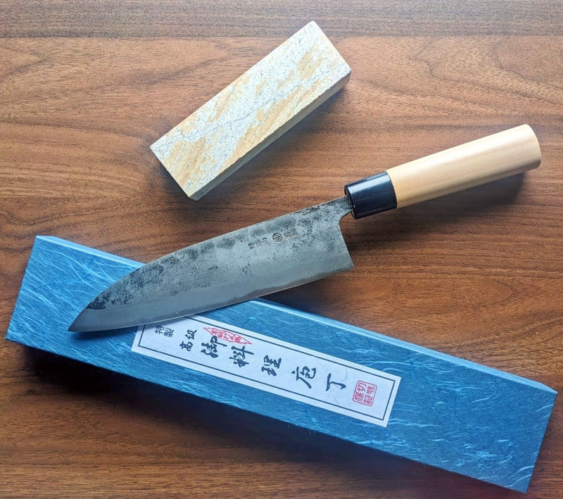 Kurouchi Deba Knife 21cm by Nakamura Hamono next to the closed box with Japanese label and the free red sharpening stone