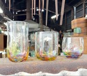 斑点のある木製の表面に副島硝子器による 3 つの手作りのレインボー グラス、背景にワークショップ