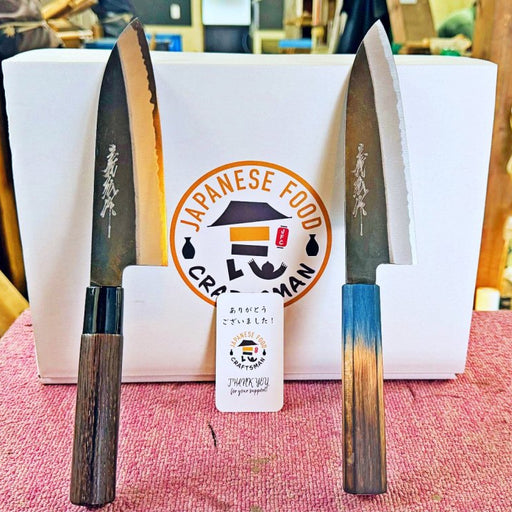 Two Yamawaki Hamono Kurouchi Santoku Knives 180mm on red table leaning on white box in workshop