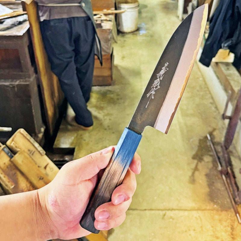 Hand holding Kurouchi Santoku Knife 180mm by Yamawaki Hamono with blue and brown handle in workshop