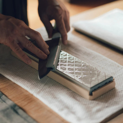 Closeup of hands sharpening a KISEKI knife with a KISEKI Knife Attachment for Sharpening on a KISEKI Diamond Whetstone in a kitchen table