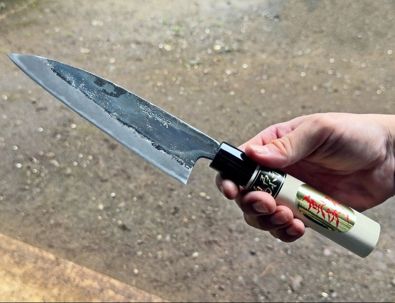 Hand holding Thin Kurouchi Deba Knife 155mm by Shigemitsu Hamono with concrete background