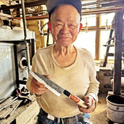 Toshio Nakamura smiling while holding a handcrafted Japanese kitchen knife in his workshop