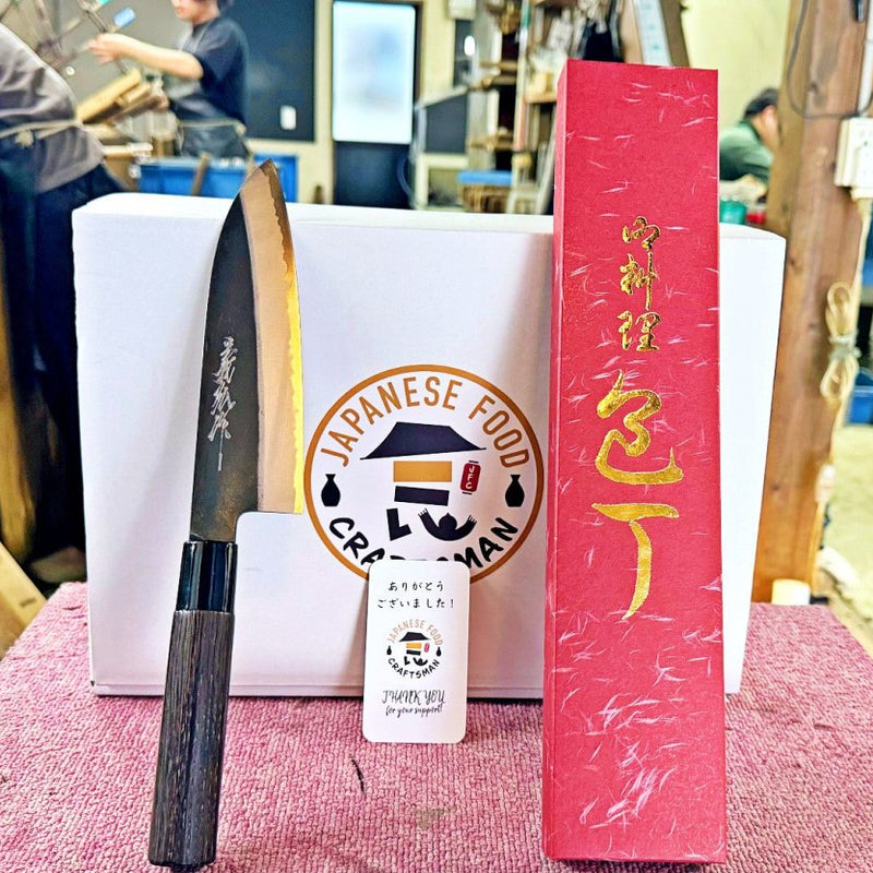 Kurouchi Santoku Knife 180mm by Yamawaki Hamono with dark brown handle on red table leaning on white box with red box