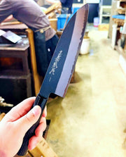 Hand holding Kurouchi Santoku Knife 180mm by Yamawaki Hamono with dark brown handle in workshop