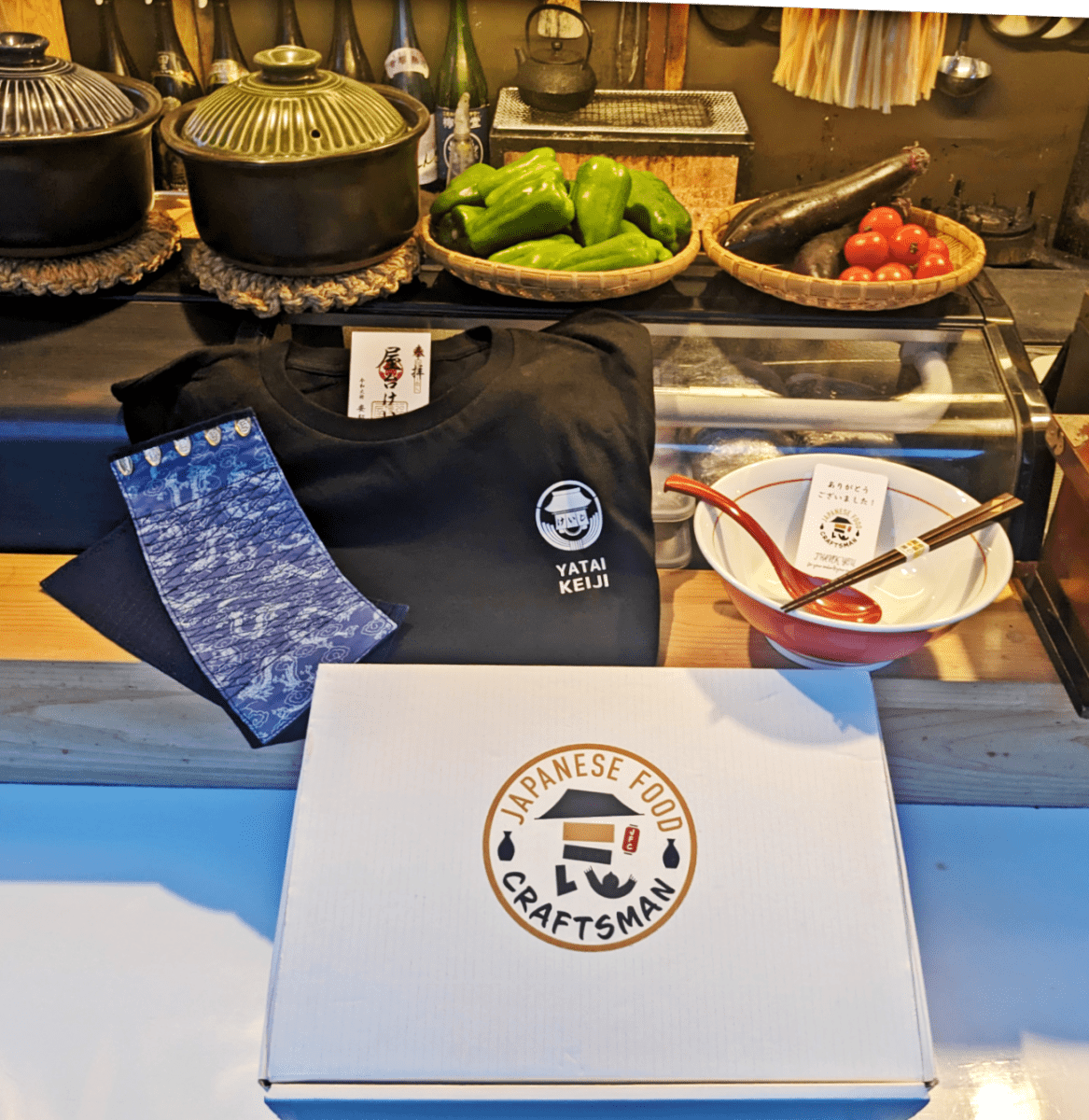 Yatai Keiji Fan Box display with blue wristbands, black Yatai Keiji t-shirt and ramen bowl set inside Yatai Keiji stall